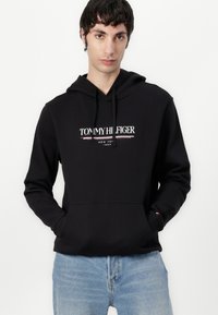 Svart hoodie med en framficka, med vit och röd text som säger "TOMMY HILFIGER NEW YORK 1985." Bärs över ljusblå jeans.