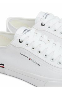 Zapatillas de lona blancas con puntera redonda, cordones planos y ojales de metal. Presentan un logotipo en el lateral y una suela de goma con acentos de color.
