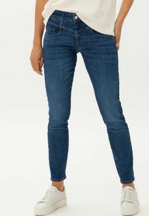 Jeans skinny en denim bleu foncé, avec une taille mi-haute, un design classique à cinq poches et un léger délavage. Portés avec des baskets blanches.