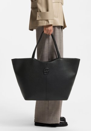Personne portant une veste beige et un pantalon gris tenant un grand sac fourre-tout en cuir noir avec des poignées doubles et un logo embossé.