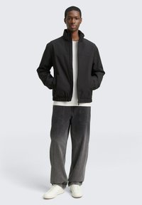 Młody mężczyzna stoi z rękami w kieszeniach, ubrany w czarną kurtkę zapinaną na zamek, białą koszulę, luźne szare dżinsy oraz białe sneakersy na tle jednolitego tła.