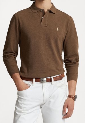 Braunes Poloshirt mit langen Ärmeln und Kragen, ausgestattet mit zwei Knöpfen und einem kleinen gestickten Logo. Kombiniert mit weißen Hosen und einem braunen Gürtel.