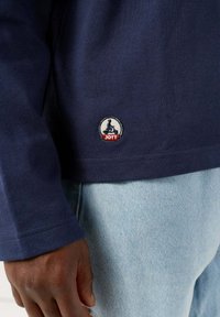 Marineblauwe longsleeve shirt van katoen met een cirkelvormig geborduurd logo met "JOTT" aan de zoom. Lichte spijkerbroek zichtbaar.