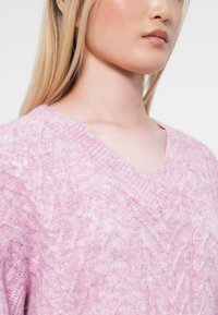 Hellrosa gestrickter Pullover mit V-Ausschnitt, strukturierten Kabelmustern und gerippten Kanten, der ein weiches und gemütliches Erscheinungsbild zeigt.
