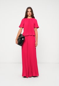 Fuksia kaittu paita ja leveälahkeiset housut, joissa on liehuva hihasuunnittelu. Malli pitää mustaa clutch-laukkua. Tausta on yksinkertainen valkoinen.