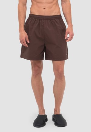 Männliches Model trägt braune Shorts mit elastischem Bund und schwarze Slip-On-Schuhe, steht vor einem schlichten hellgrauen Hintergrund.