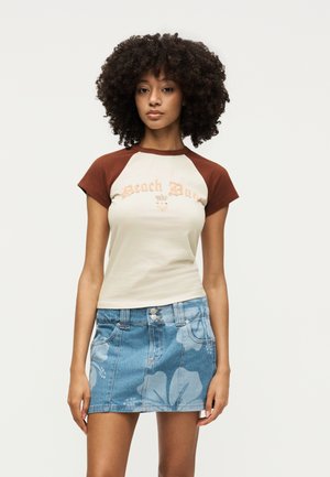 Jeune femme aux cheveux bouclés portant un t-shirt beige et marron « Beach Bum » et une mini-jupe en jean bleu à fleurs, debout devant un fond uni.