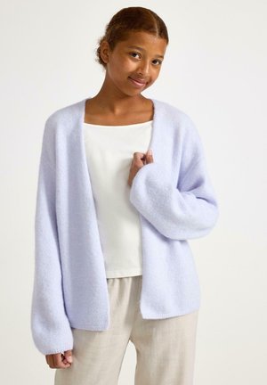 Junge Person mit lockigem Haar trägt eine weiche, hellviolette Strickjacke über einem weißen Oberteil und beigen Hosen, stehend vor einem einfarbigen Hintergrund.