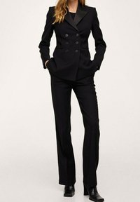 Costume noir sur mesure avec un blazer croisé, des revers à cran et des poches avant. Pantalon ajusté à coupe droite et texture lisse.