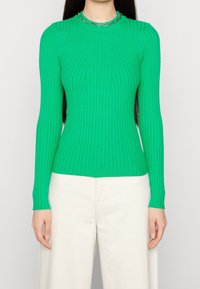 Maglione verde a maniche lunghe con collo alto e motivo a coste. Abbinato a pantaloni chiari a gamba larga.