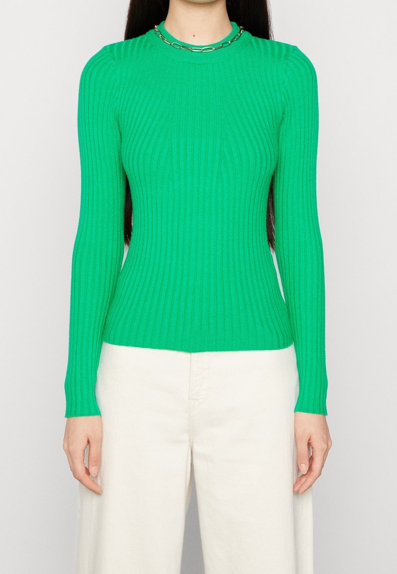 Maglione verde a maniche lunghe con collo alto e motivo a coste. Abbinato a pantaloni chiari a gamba larga.