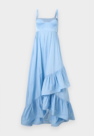 Ženska v svetlo modri asimetrični obleki z volančki, bustier zgornji del in srebrnimi sandali, stoji pred enobarvnim belim ozadjem.