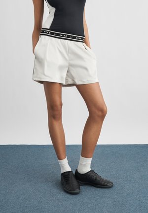 Person iført hvide Nike-shorts med sort logo-bånd, en sort og hvid top, hvide sokker og sorte sko, der står på blåt tæppe.
