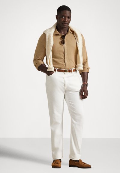 Vyras vilki smėlio spalvos marškinius su sagomis, baltas kelnes, ruda diržu ir batais, ant pečių perdetas kreminis megztinis, nuo apykaklės kabančias saulės akinius.