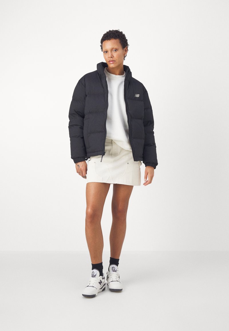 Schwarze Daunenjacke mit hohem Kragen, Reißverschluss und Logodetail; getragen über einem weißen Pullover und einem beigen Rock, kombiniert mit weißen Sneakers.