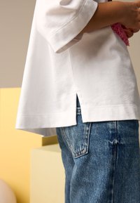 White cotton t-shirt with short sleeves, featuring a side split at the hem, paired with blue denim jeans showing faded texture and back pockets.