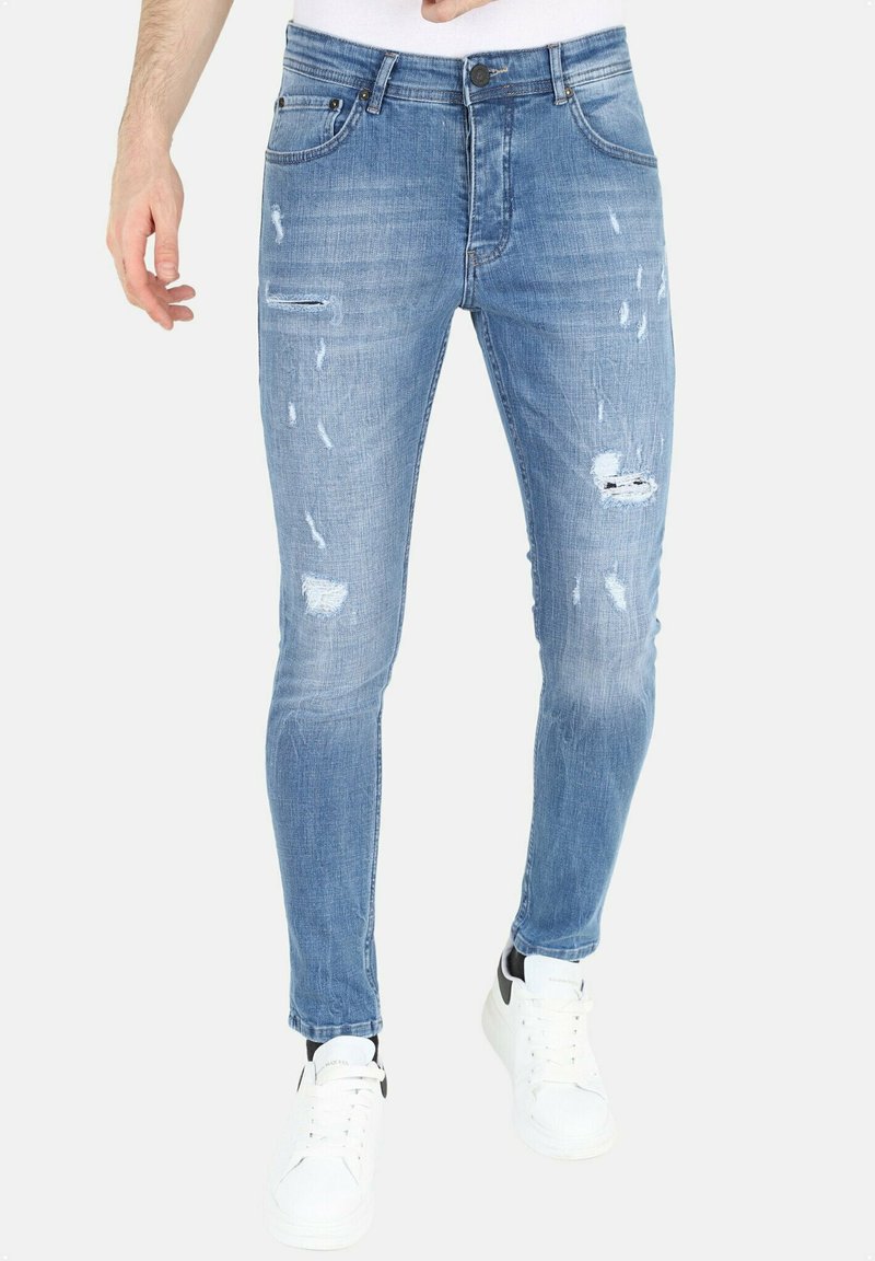 Lichtblauwe skinny jeans met verwerkte details, waaronder scheuren en vervaging, gecombineerd met witte sneakers met zwarte accenten.