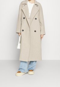 Beige double-breasted jas van een wolmix met een brede kraag, zwarte knopen en een ontspannen pasvorm, gecombineerd met lichtblauwe jeans met wijd uitlopende pijpen en beige plateauzolen.