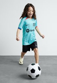 Un bambino indossa una maglia da calcio del Barcellona di colore teal con accenti rossi e blu mentre dribbla un pallone da calcio bianco e nero, abbinato a pantaloni neri.