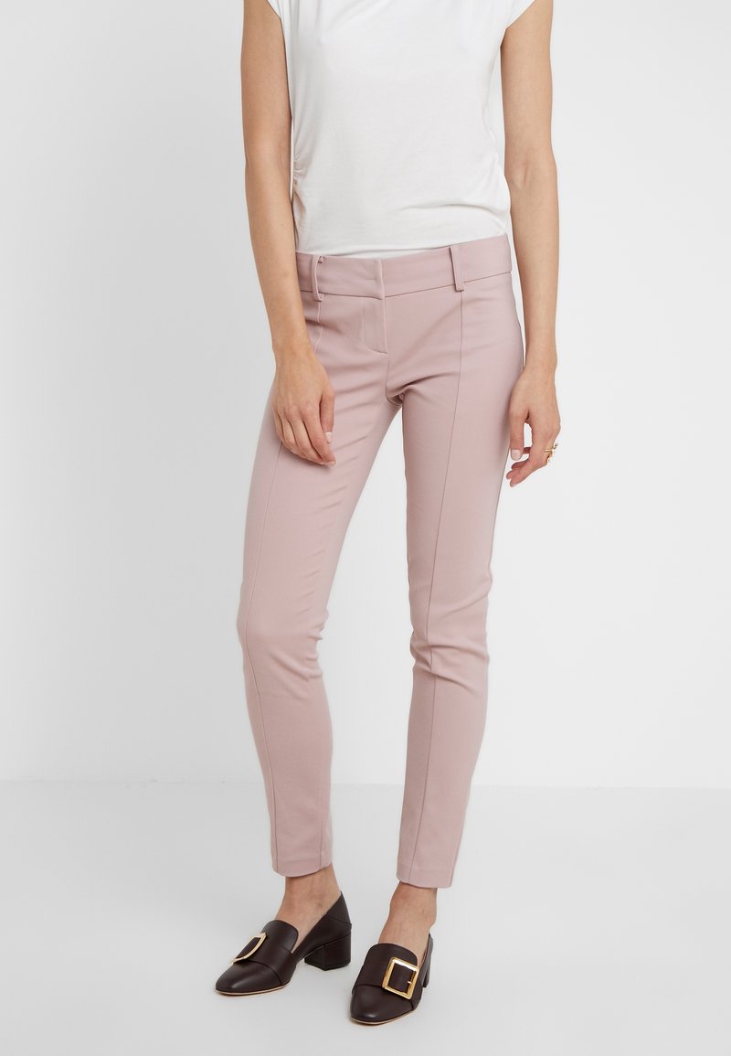 Vrouw in licht mauve, strakke broek met zwarte blokhak-schoenen met grote gouden gespen, tegen een effen witte achtergrond.