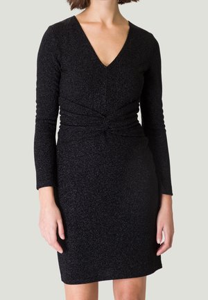 Vrouw draagt een nauwsluitende zwarte jurk met lange mouwen, een V-hals en een gedraaid detail ter hoogte van de taille tegen een effen lichte achtergrond.