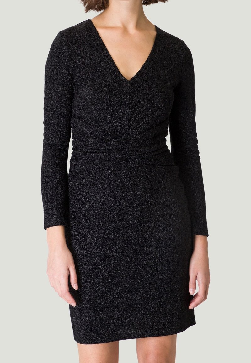 Femme portant une robe noire ajustée à manches longues avec un col en V et un détail torsadé à la taille sur un fond clair uni.