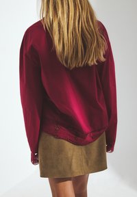 Vrouw met lang blond haar, gekleed in een losvallende rode blouse met kant aan de onderkant en een tan suède minirok, staand tegen een effen witte achtergrond.