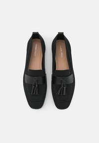 Zapatos negros sin cordones con parte superior de punto texturizado, que presentan una banda negra lisa con acentos de borlas. El interior tiene un forro beige.