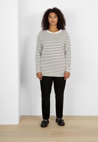 Dlhý rukáv, pruhovaný top v off-white a čiernej farbe. Spárovaný s čiernymi nohavicami rovného strihu. Nosené s hrubými čiernymi topánkami. Pozadie zo snehobieleho dreveného podlahy.