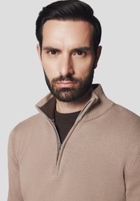 Beżowy sweter z zapięciem na zamek, wykonany z teksturowanej dzianiny, z wysokim kołnierzem i kontrastującą ciemniejszą wewnętrzną warstwą. Charakteryzuje się minimalistycznym wzornictwem.