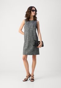 Grijs mouwloos jurkje van een textuurstof, gecombineerd met zwarte sandaaltjes en een zwarte clutch. Haar in losse krullen gestyled.