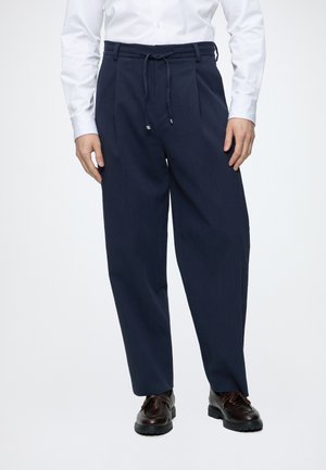 Homme portant un pantalon plissé bleu marine avec taille à cordon, chemise blanche et chaussures en cuir marron foncé, debout devant un fond blanc.