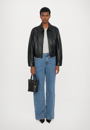 Schwarze Lederjacke in Cropped-Optik über einem hellgrauen Oberteil, kombiniert mit hochtaillierten blauen Schlaghosen und einer kleinen schwarzen Handtasche.