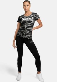 Schwarz-graues T-Shirt mit Camouflage-Muster und Logo, kombiniert mit schwarzen Leggings mit seitlichem Camouflage-Streifen, ergänzt durch weiße Sneakers.