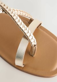 Metalen gouden gevlochten bandjes op een beige platte sandaal met een glad, textured voetbed en een eenvoudige metalen accent op de bovenste band.