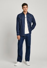 Donkerblauwe denim shirt met lichte stiksels, twee borstzakken, gelaagd over een wit t-shirt; gecombineerd met bijpassende donkerblauwe jeans en witte sneakers.