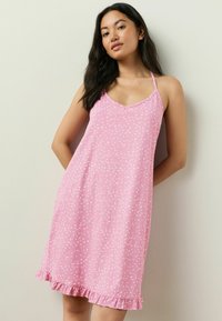 Ung kvinde med langt mørkt hår iført en pink ærmeløs kjole med hvide prikker og flæse, stående mod en lys væg.