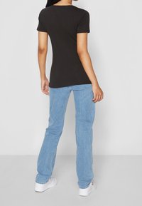 En person står vänd bort från kameran i en åtsittande svart t-shirt och ljusblå jeans mot en neutral ljus bakgrund. Vita sneakers syns.