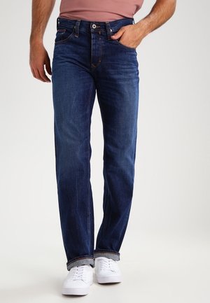 Mężczyzna w ciemnoniebieskich dżinsach z podwiniętymi nogawkami, w wpuszczonej w spodnie różowawej koszuli i białych trampkach, stoi na jednolitym tle.