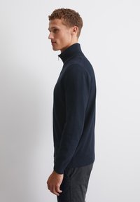 Marine gebreide pullover met rits, voorzien van een textuurafwerking, een getailleerde pasvorm en ribgebreide manchetten. Gepaarde met donkere broek, zijaanzicht getoond.