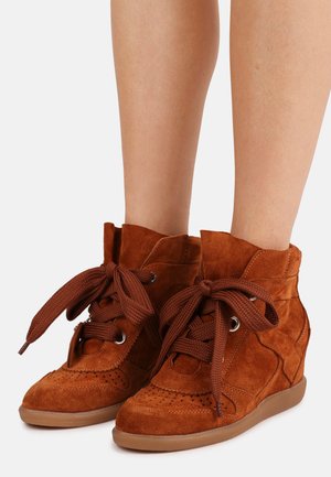 Καφέ σουέτ ψηλοτά αθλητικά παπούτσια με μεγάλα πλεχτά κορδόνια, διάτρητες λεπτομέρειες και λαστιχένιες σόλες, φορεμένα σε γυμνά πόδια πάνω σε λευκό φόντο.