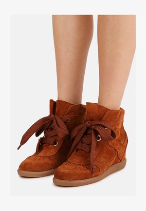 Braune Keilsneaker aus Wildleder mit großen geflochtenen Schnürsenkeln, perforierten Details und Gummisohlen, getragen an nackten Beinen vor weißem Hintergrund.