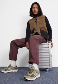 Chaleco de forro polar marrón con detalles en negro, camisa negra de manga larga, pantalones burdeos, calcetines blancos, zapatillas verdes y grises, sentado sobre una caja plateada.