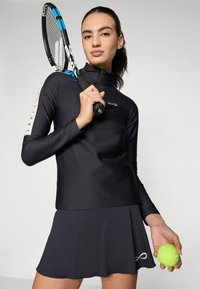 Schwarzes langärmliges Sportoberteil mit Reißverschlusskragen, kombiniert mit einem schwarzen ausgestellten Rock. Hält einen Tennisschläger und einen leuchtend grünen Tennisball.