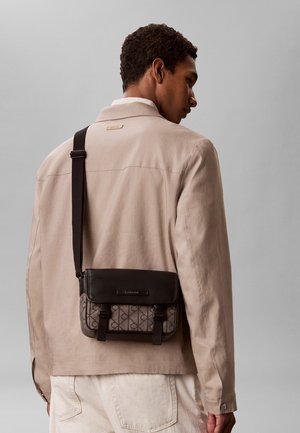 Mężczyzna w beżowej kurtce i kremowych spodniach niesie brązową, wzorzystą torbę na ramię z czarną klapką i paskiem.