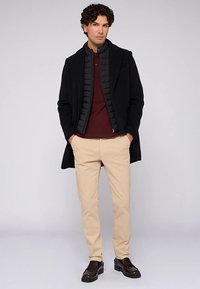 Schwarzer Mantel über einer grauen Steppweste, bordeauxrotem Poloshirt, beigen Hosen und dunkelbraunen Schuhen. Glatter Stoff mit tailliertem Schnitt.