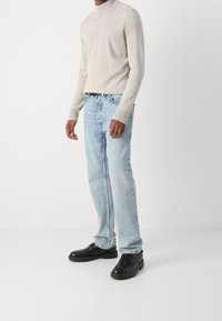 Pull à col roulé en maille gris clair, jean en denim bleu et chaussures noires à lacets. Le jean présente une coupe décontractée et une légère usure.