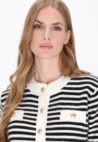 Zwart-wit gestreepte cardigan met goudkleurige knopen, ronde halslijn en textuurstof. Beschikt over twee borstzakken met vergelijkbare knopdetails.