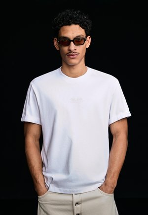 Mladić s tamnom kovrčavom kosom i brkovima, nosi tamne pravokutne sunčane naočale, bijelu majicu i bež hlače s rukama u džepovima.