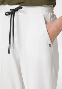 Pantalones de chándal blancos con cintura elástica, cordón negro y bolsillos laterales. Presenta un pequeño logo negro cerca del bolsillo. Textura suave.